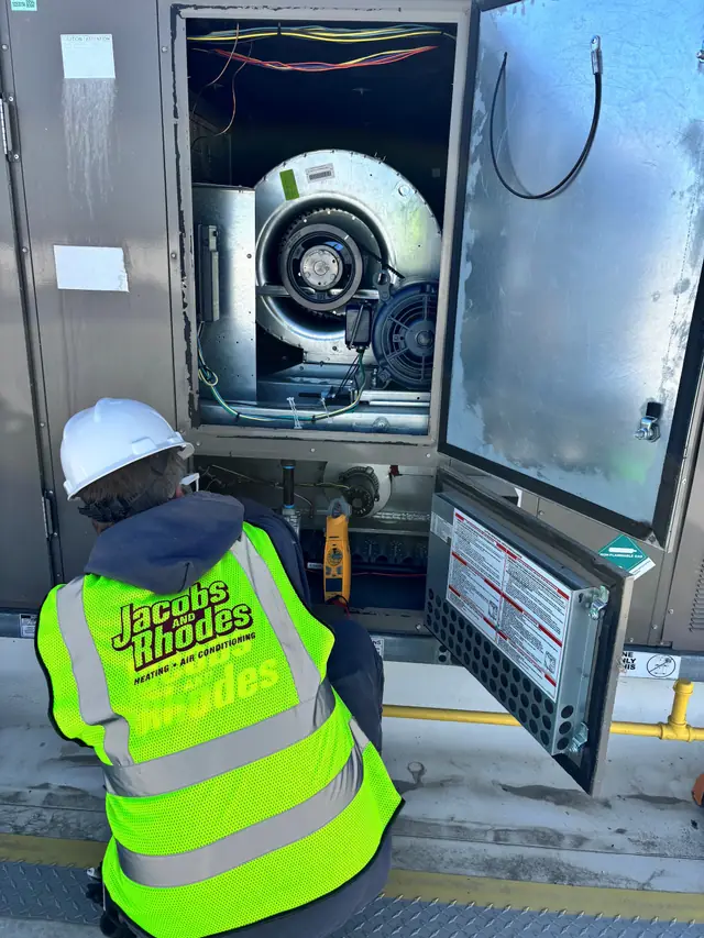 A Jacobs and Rhodes technician in a white hard hat and hi-vis vest servicing a rooftop unit with the blower and burner section open